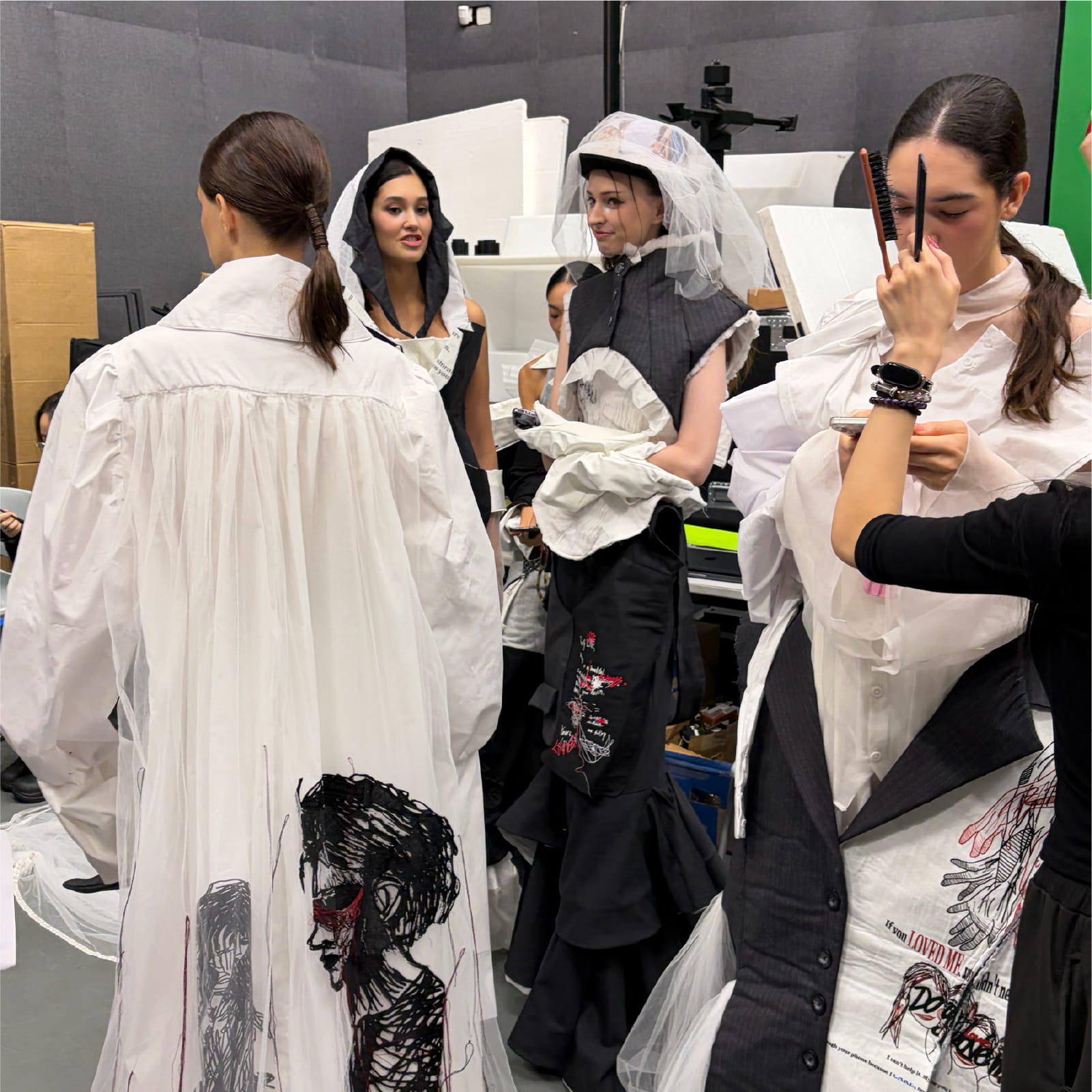 Backstage view of models preparing for the runway, highlighting the intricate black and red facial embroidery on the back of a flowing white garment.