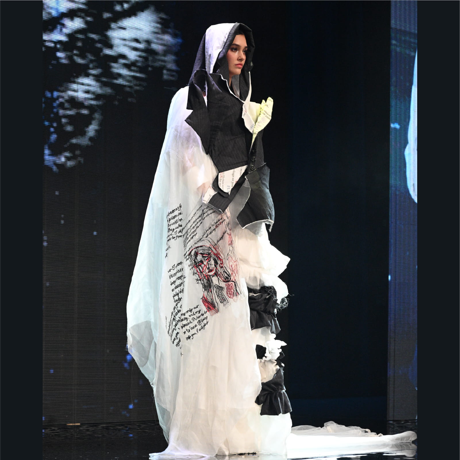 A runway model showcasing a layered black and white outfit with a prominent red and black embroidered face design on the skirt, from Yuen Wing Ki's collection.