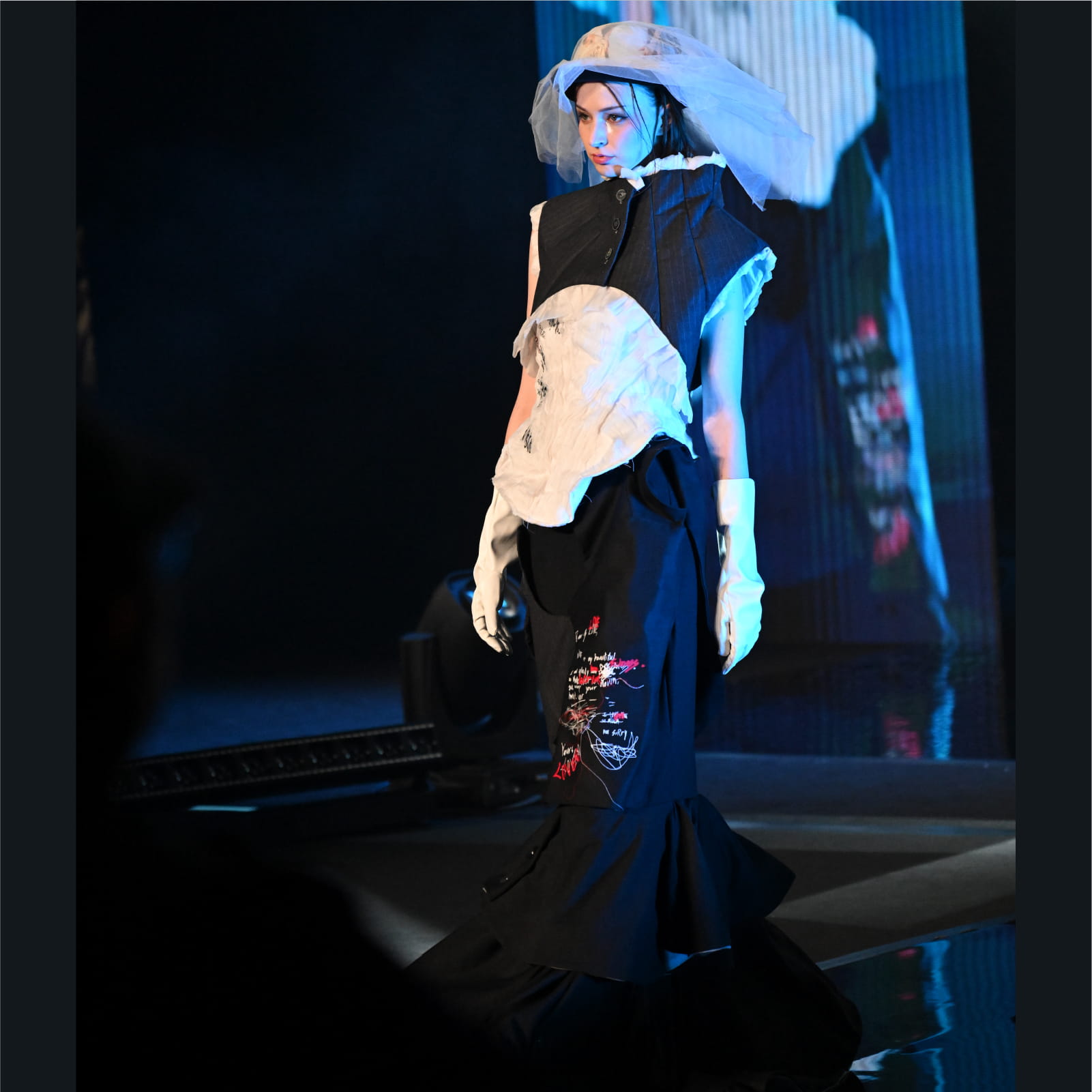 A model walks the runway in a structured black and white outfit featuring striking red and white embroidered text and abstract lines on the dark skirt.