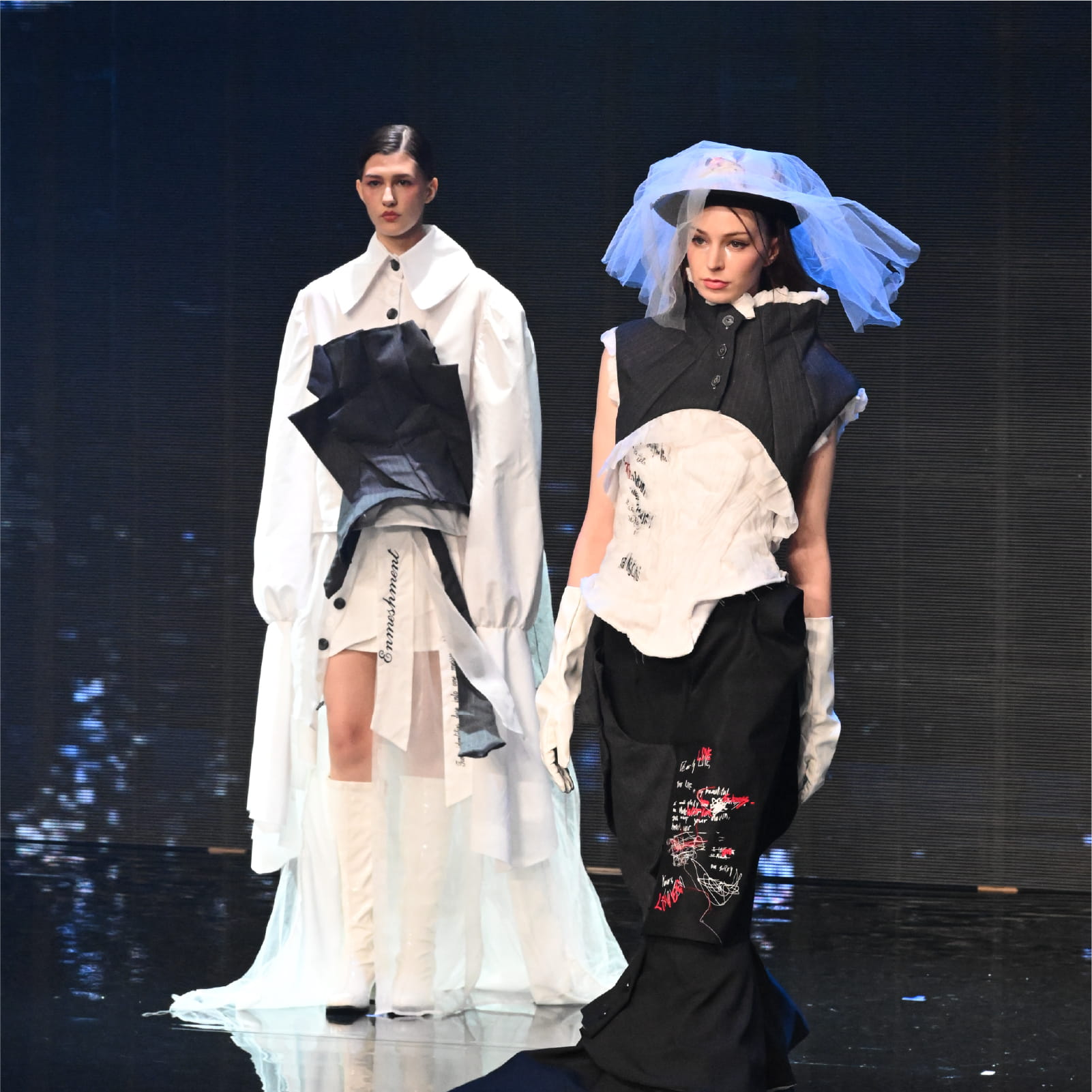 Two models presenting deconstructed black and white suits and dresses from the "PATHOLOGICAL LOVE" collection on the HKDI runway, showcasing a mix of structured tailoring and flowing fabrics.